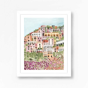 May include: Watercolor painting of a hillside town with buildings in shades of pink, peach, and white. Green foliage and purple flowers are in the foreground. The sky is a light blue. The artwork is framed in a white border.