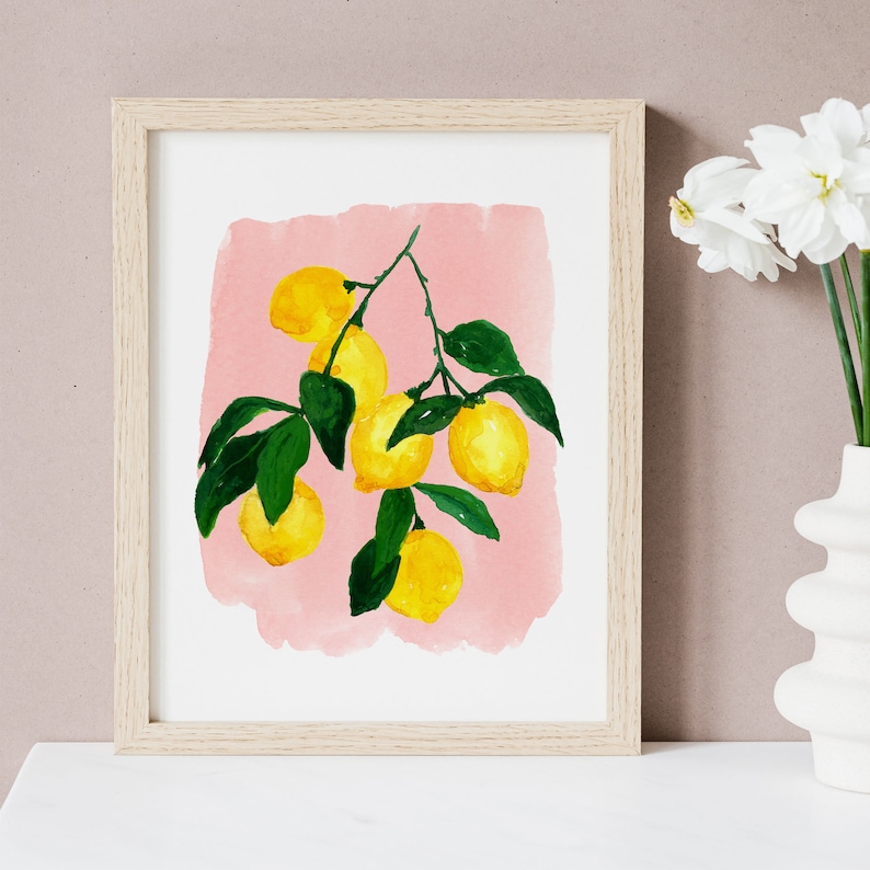 May include: A framed watercolor print featuring a branch of lemons with green leaves against a pink background. The lemons are yellow, and the frame is light wood. The artwork is displayed on a white surface.