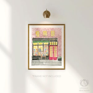 May include: Watercolor print of a bookstore with a pink brick facade, green awning, and bright pink doors. The word "BOOKS" is above the entrance. A bench and table with flowers are in front of the store. The print is in a gold frame.