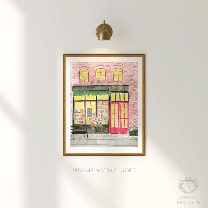 May include: Watercolor print of a bookstore with a pink brick facade, green awning, and bright pink doors. The word "BOOKS" is above the entrance. A bench and table with flowers are in front of the store. The print is in a gold frame.