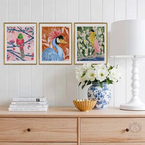 May include: Three framed watercolor bird prints with gold frames. The artwork features colorful birds on branches and in floral settings. The prints are displayed on a wooden cabinet with a lamp, vase, and books.