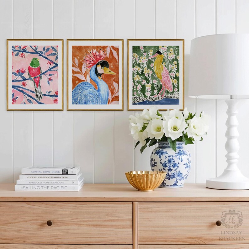 May include: Three framed watercolor bird prints with gold frames. The artwork features colorful birds on branches and in floral settings. The prints are displayed on a wooden cabinet with a lamp, vase, and books.