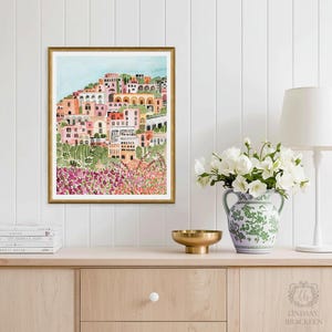 May include: A framed watercolor print of a hillside village with colorful buildings in shades of pink, orange, and white. The artwork is set against a light blue sky and framed in gold. The print is displayed on a wooden cabinet with a vase of white flowers.