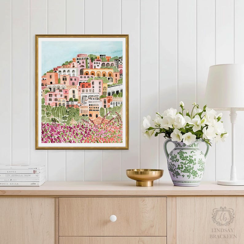 May include: A framed watercolor print of a hillside village with colorful buildings in shades of pink, orange, and white. The artwork is set against a light blue sky and framed in gold. The print is displayed on a wooden cabinet with a vase of white flowers.