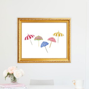 May include: A framed print featuring watercolor illustrations of five beach umbrellas in various colors and patterns, including red and white stripes, green and yellow stripes, and blue and white stripes. The frame is gold.