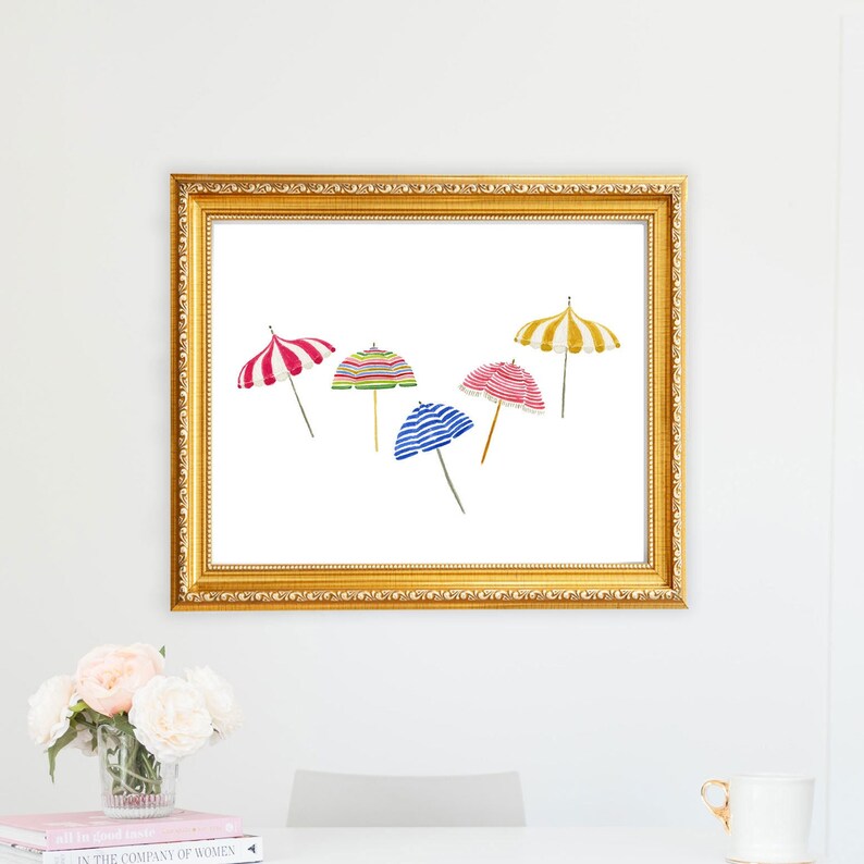 May include: A framed print featuring watercolor illustrations of five beach umbrellas in various colors and patterns, including red and white stripes, green and yellow stripes, and blue and white stripes. The frame is gold.