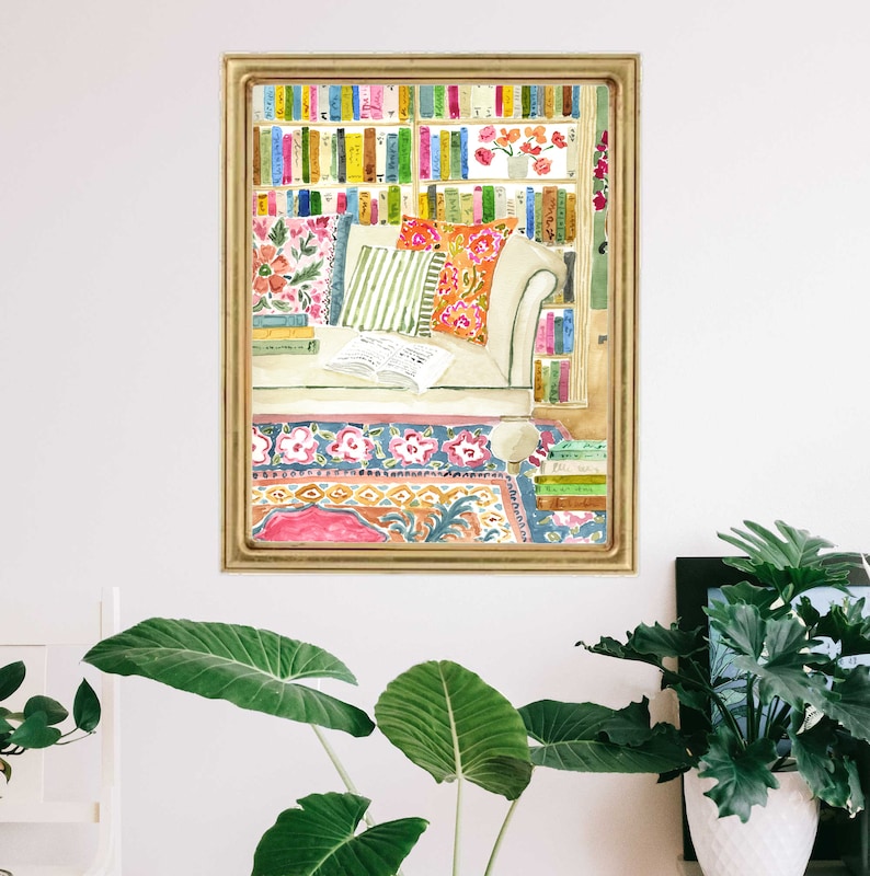 May include: Watercolour painting of a reading nook. A white sofa with patterned cushions sits before a bookshelf filled with colourful books. A patterned rug and open book complete the scene, framed in a gold border.