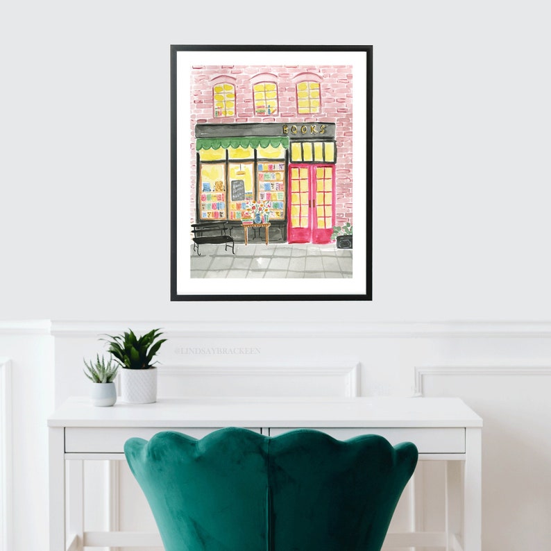 May include: Watercolor painting of a bookstore exterior. The building has a pink brick facade, a black awning, and a bright red door. The word "BOOKS" is above the entrance. A bench and table with flowers are in front of the store.