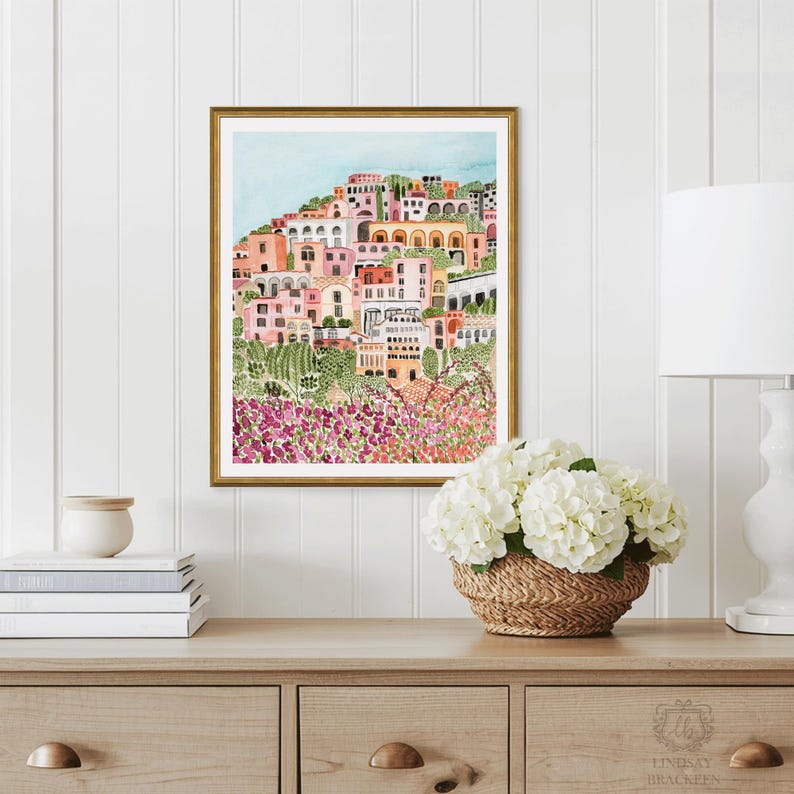May include: Watercolor print of a coastal Italian town in a gold frame. The artwork depicts colorful buildings on a hillside, with pink and purple flowers in the foreground. The sky is light blue.