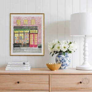 May include: A framed watercolor print of a bookstore with a pink brick facade, red doors, and a green awning. The print is displayed on a wooden cabinet with books, a gold bowl, a blue and white vase, and a white lamp.