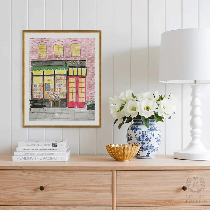 May include: A framed watercolor print of a bookstore with a pink brick facade, red doors, and a green awning. The print is displayed on a wooden cabinet with books, a gold bowl, a blue and white vase, and a white lamp.