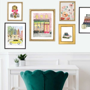 May include: A collection of framed watercolor art prints. The prints feature various subjects, including a New York City street scene with a yellow taxi, a bookstore facade, and floral arrangements. The frames are gold and black, and the overall aesthetic is artistic and decorative.