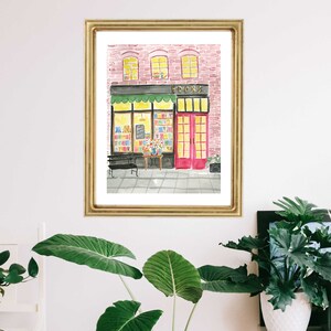 May include: Watercolor painting of a bookstore exterior in a gold frame. The building has a pink brick facade, a green awning, and a red door. The windows are lit, and the word "BOOKS" is above the entrance.