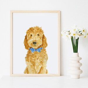 May include: A framed watercolor painting of a golden-brown curly-haired dog wearing a bright blue bow tie. The dog is depicted from the chest up, facing forward with a neutral expression, set against a plain white background within a light wood frame.