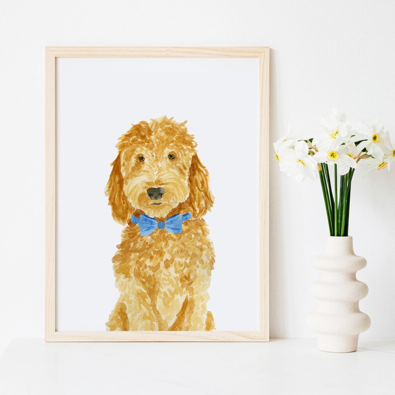 May include: A framed watercolor painting of a golden-brown curly-haired dog wearing a bright blue bow tie. The dog is depicted from the chest up, facing forward with a neutral expression, set against a plain white background within a light wood frame.