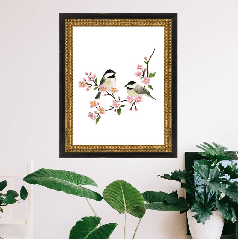May include: A framed watercolour print depicting two small birds on a flowering branch with pink blossoms and green leaves. The artwork is set against a white background and framed in a black and gold ornate frame, suitable for home decor.