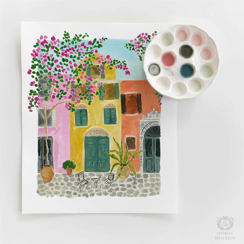 May include: A watercolor painting of a European street scene with pink, yellow, and orange buildings, a cobblestone ground, and a flowering tree. A white circular paint palette with various colors sits in the upper right corner of the artwork.