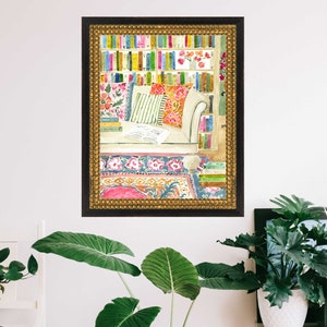 May include: Watercolour painting of a reading corner. A white sofa with colourful cushions is in front of a bookshelf filled with books. A patterned rug and a gold framed border complete the scene. The artwork is displayed on a white wall.