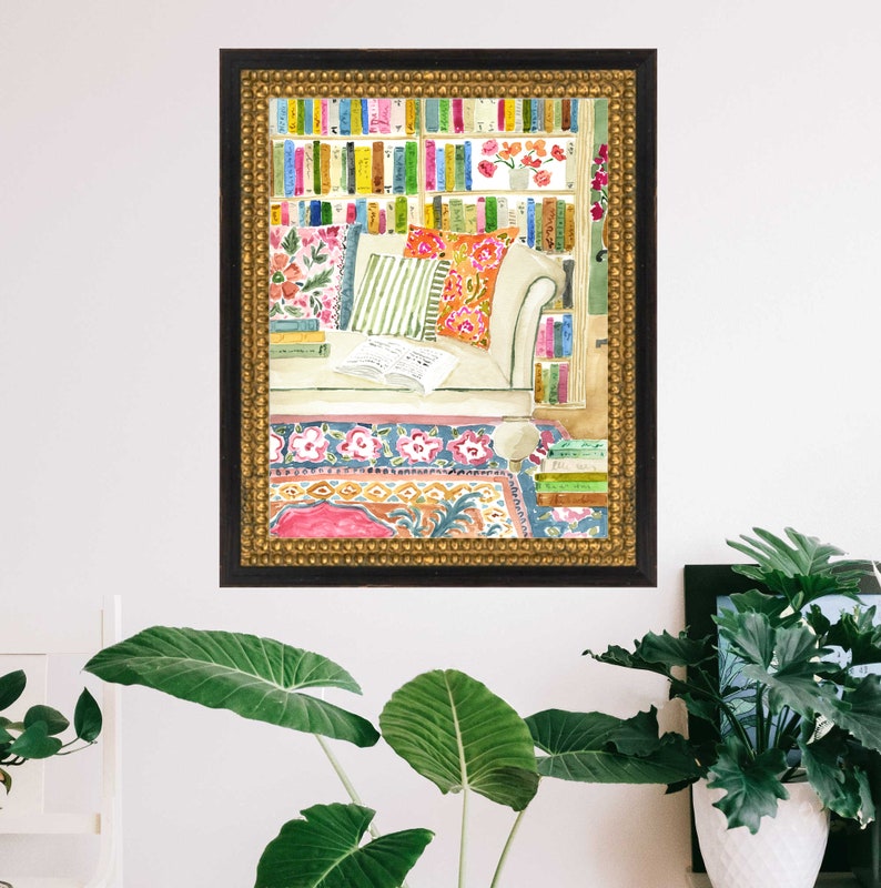 May include: Watercolour painting of a reading corner. A white sofa with colourful cushions is in front of a bookshelf filled with books. A patterned rug and a gold framed border complete the scene. The artwork is displayed on a white wall.