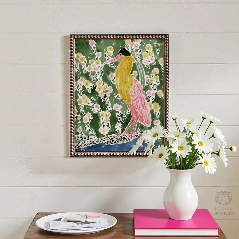 May include: A framed watercolor painting of a bird with yellow and pink plumage, set against a green floral background. The frame is brown with a beaded edge. A white vase with daisies and a pink book are on a wooden table.
