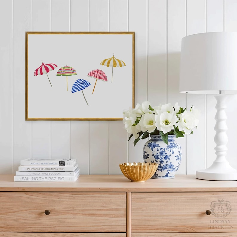 May include: A framed print featuring watercolor illustrations of five beach umbrellas in various colors, including red, yellow, and blue. The artwork is displayed above a wooden dresser with books, a gold bowl, and a floral arrangement.