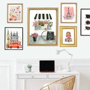 May include: A white desk with a gold metal chair, a laptop, a lamp, and a potted plant. There are eight framed prints on the wall above the desk, including images of flowers, the Eiffel Tower, Big Ben, and a red double-decker bus. The prints are all in gold frames.