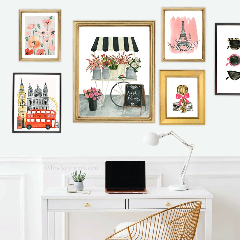 May include: A white desk with a gold metal chair, a laptop, a lamp, and a potted plant. There are eight framed prints on the wall above the desk, including images of flowers, the Eiffel Tower, Big Ben, and a red double-decker bus. The prints are all in gold frames.