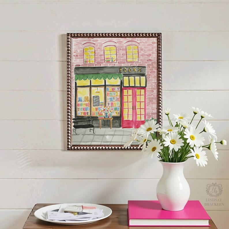 May include: Watercolor painting of a bookstore with a green awning, red door, and the word "BOOKS" above the entrance. The painting is in a decorative frame. A white vase with daisies sits on a table with a pink book.