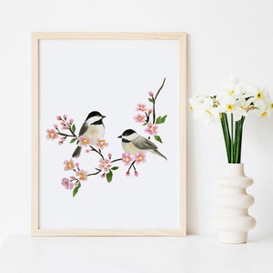 May include: Watercolour print featuring two small birds on a flowering branch with pink blossoms. The birds have black and white markings. The artwork is framed in a light wood frame and is displayed on a white surface.