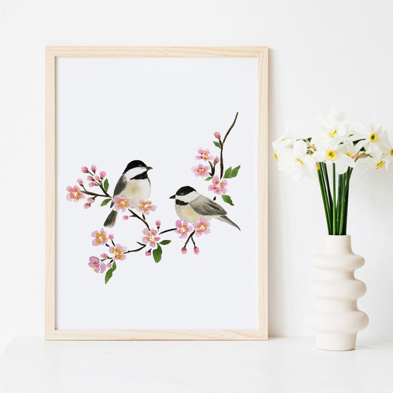 May include: Watercolour print featuring two small birds on a flowering branch with pink blossoms. The birds have black and white markings. The artwork is framed in a light wood frame and is displayed on a white surface.
