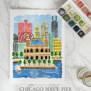 May include: A watercolor art print of the Chicago skyline featuring Navy Pier, a Ferris wheel, and various buildings in shades of pink, yellow, and blue. The print includes the text CHICAGO NAVY PIER ART PRINT at the bottom, with a paint set nearby.
