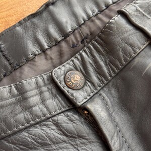 Pu&ograve; includere: Primo piano di pantaloni in pelle grigio scuro. I pantaloni presentano una chiusura con bottone con un bottone circolare con la scritta "DIESEL". La pelle ha un aspetto strutturato e cuciture visibili.