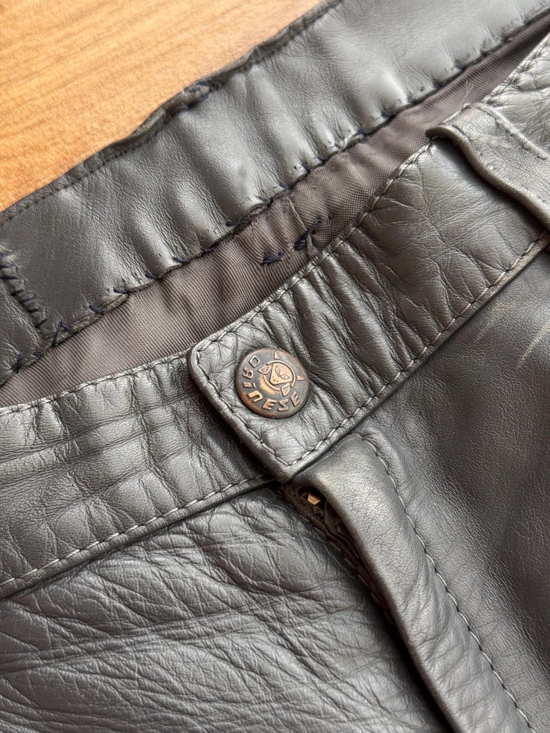 Pu&ograve; includere: Primo piano di pantaloni in pelle grigio scuro. I pantaloni presentano una chiusura con bottone con un bottone circolare con la scritta "DIESEL". La pelle ha un aspetto strutturato e cuciture visibili.