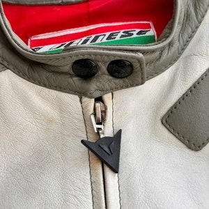Op de afbeelding: Een close-up van een wit leren jack met een grijze kraag en een zwarte rits. Het jack heeft een Dainese-logo op de rits en een rood-groen Dainese-label aan de binnenkant van de kraag.