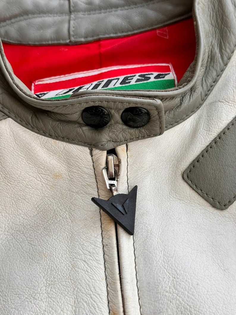 Op de afbeelding: Een close-up van een wit leren jack met een grijze kraag en een zwarte rits. Het jack heeft een Dainese-logo op de rits en een rood-groen Dainese-label aan de binnenkant van de kraag.