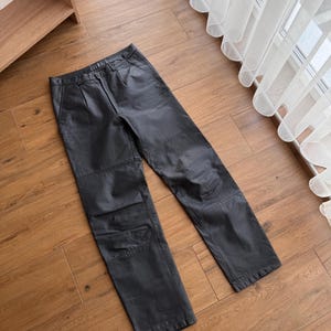 Pu&ograve; includere: Pantaloni in pelle nera con un classico design a gamba dritta. I pantaloni presentano una chiusura con bottone e cerniera, passanti per cintura e tasche. La pelle sembra morbida e ha una leggera lucentezza. I pantaloni sono appoggiati su una superficie di legno.