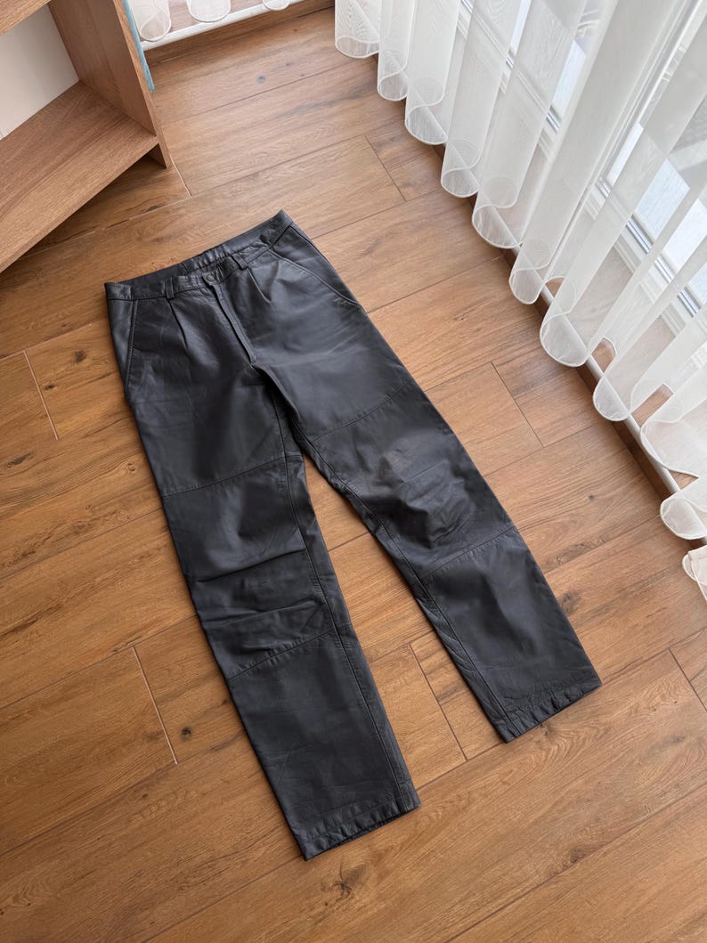Pu&ograve; includere: Pantaloni in pelle nera con un classico design a gamba dritta. I pantaloni presentano una chiusura con bottone e cerniera, passanti per cintura e tasche. La pelle sembra morbida e ha una leggera lucentezza. I pantaloni sono appoggiati su una superficie di legno.