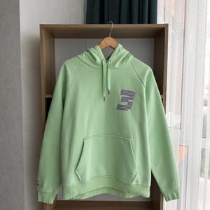 Può includere: Una felpa con cappuccio verde chiaro con una grande lettera "E" stilizzata stampata sul davanti. La felpa ha una tasca a marsupio e un cappuccio con coulisse.