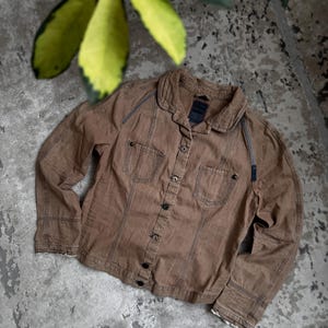 May include: A brown button-down shirt with two chest pockets and a collar. The shirt has long sleeves and is made of a textured fabric. The stitching is visible, and the shirt appears to be worn. The shirt is on a grey surface.