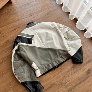 Op de afbeelding: Een wit, grijs en zwart leren jack met een geometrisch ontwerp. Het jack heeft een ritssluiting en een opstaande kraag.