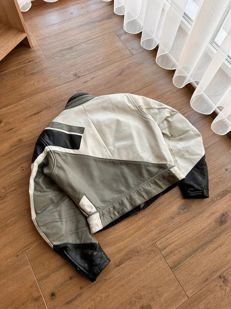 Op de afbeelding: Een wit, grijs en zwart leren jack met een geometrisch ontwerp. Het jack heeft een ritssluiting en een opstaande kraag.