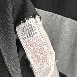 Op de afbeelding: Een zwart en grijs sweatshirt met een wit label waarop "Gemaakt in Indonesi&euml;" staat en met wasinstructies in meerdere talen.