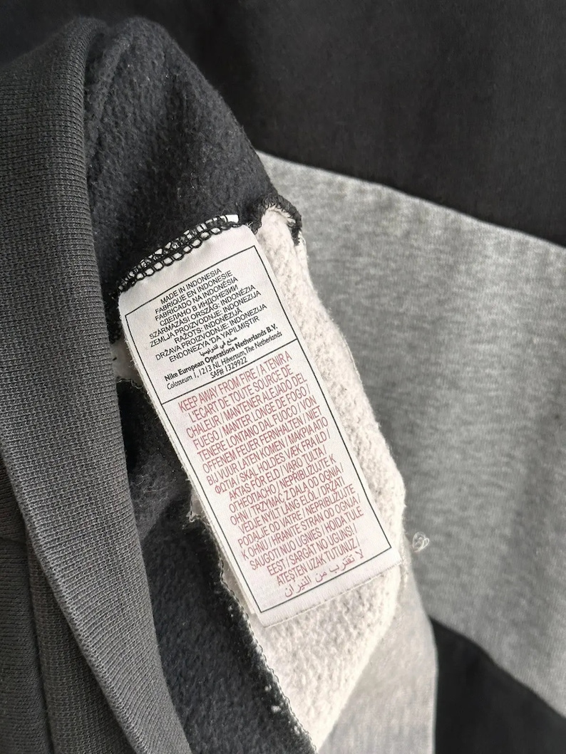 Op de afbeelding: Een zwart en grijs sweatshirt met een wit label waarop "Gemaakt in Indonesi&euml;" staat en met wasinstructies in meerdere talen.