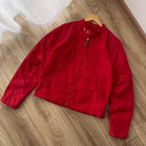 May include: A bright red jacket with a stand-up collar, a zipper closure, and two front pockets. The jacket is hanging on a wooden hanger. The jacket is made of a smooth fabric.