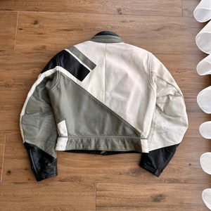 Op de afbeelding: Een wit leren jack met zwarte en grijze geometrische panelen. Het jack heeft een ritssluiting en een opstaande kraag.