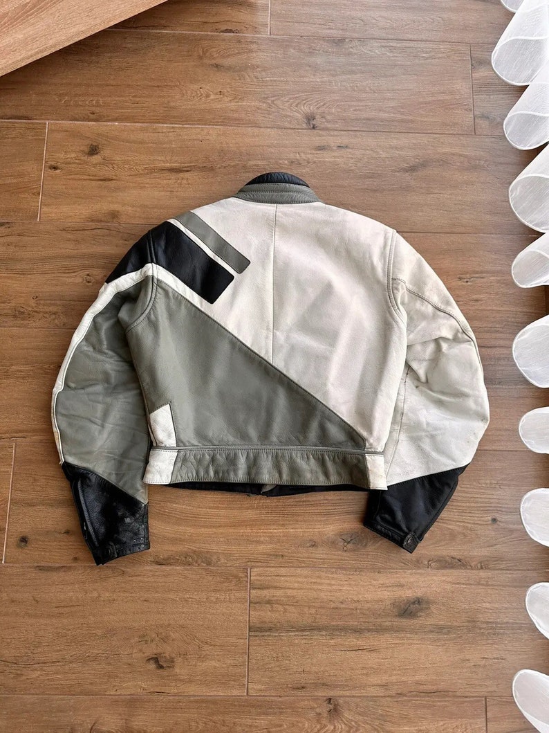 Op de afbeelding: Een wit leren jack met zwarte en grijze geometrische panelen. Het jack heeft een ritssluiting en een opstaande kraag.