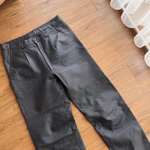 Pu&ograve; includere: Pantaloni in pelle nera con chiusura a bottone e pieghe in vita. I pantaloni hanno un classico design a cinque tasche e sono realizzati in pelle liscia e morbida. Sono appoggiati su una superficie di legno.