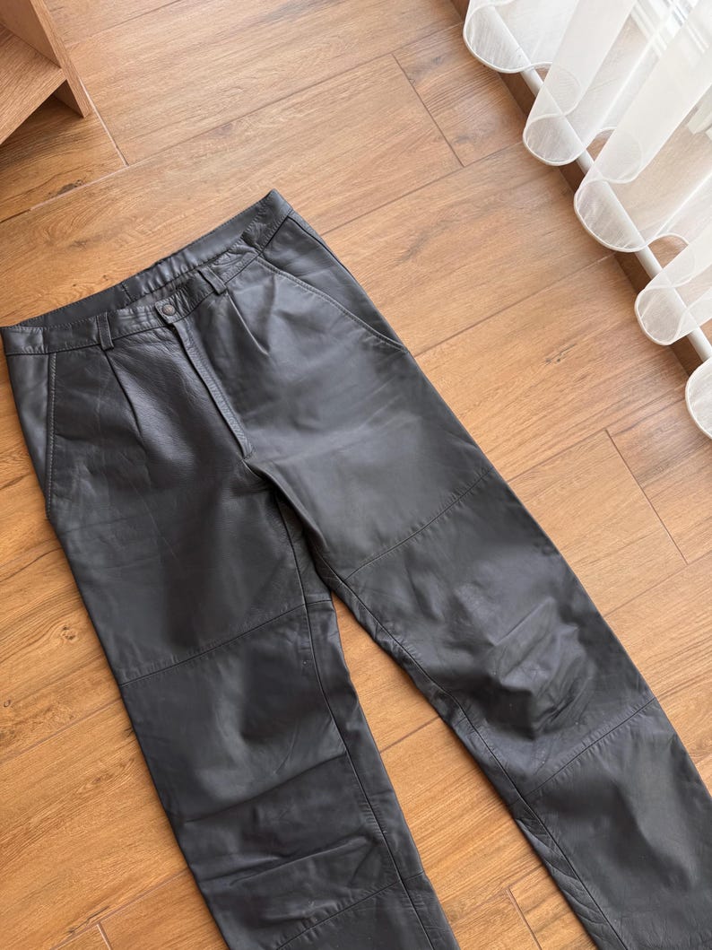Pu&ograve; includere: Pantaloni in pelle nera con chiusura a bottone e pieghe in vita. I pantaloni hanno un classico design a cinque tasche e sono realizzati in pelle liscia e morbida. Sono appoggiati su una superficie di legno.