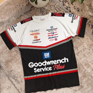 Könnte beinhalten: Weißes, schwarzes und rotes Kurzarm-T-Shirt mit mehreren Logos und Text. Das Shirt zeigt den Text "Goodwrench Service Plus" in großen weißen Buchstaben auf einem schwarzen Feld. Logos sind "GM", "ACDelco" und "Goodyear".