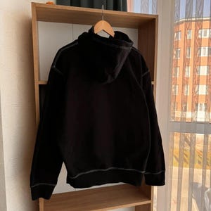 Pu&ograve; includere: Felpa con cappuccio nera con dettagli di cuciture bianche. La felpa &egrave; appesa a uno scaffale di legno.